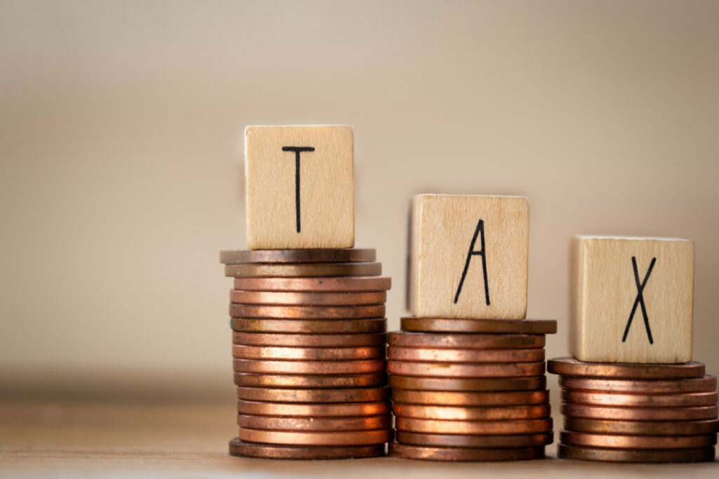Three wooden blocks that spell the word "tax" resting on coin stacks of different heights